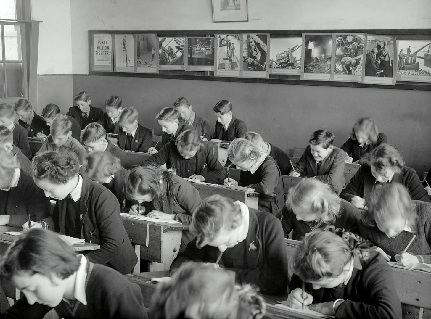 A black and white photo of a classroom in the 1940's, with rows of students bent over their work. A black and white photo of a classroom in the 1940's, with rows of students bent over their work.