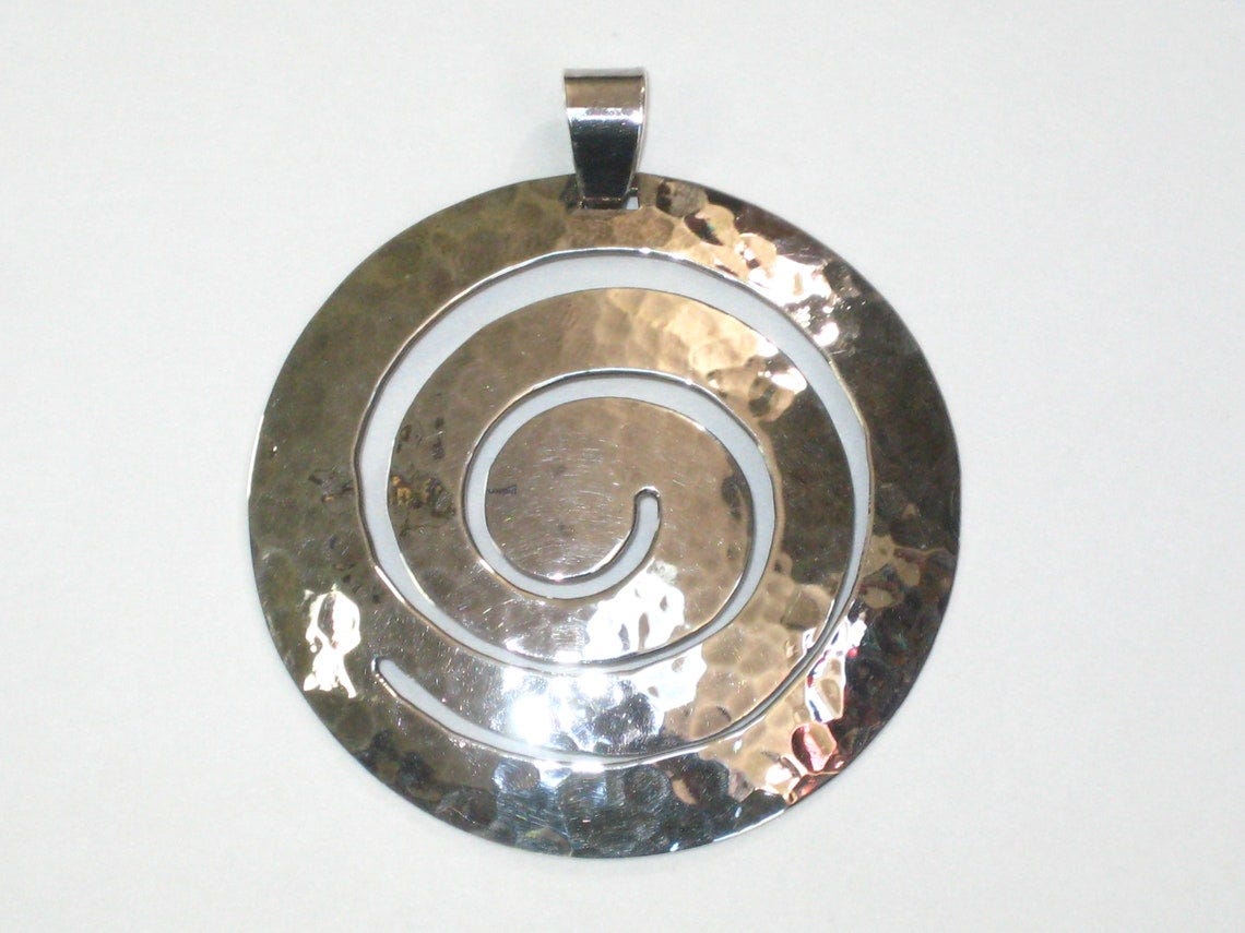May include: Silver spiral pendant. The pendant is round with a hammered texture and features a cutout spiral design. The bail is simple and understated. This modern, minimalist pendant is perfect for adding a touch of elegance to any outfit. The pendant's hammered surface adds a unique texture and dimension, while the clean lines of the spiral create a visually appealing contrast. The overall design is both sophisticated and eye-catching, making it a versatile piece for everyday wear or special occasions. May include: Silver spiral pendant. The pendant is round with a hammered texture and features a cutout spiral design. The bail is simple and understated. This modern, minimalist pendant is perfect for adding a touch of elegance to any outfit. The pendant's hammered surface adds a unique texture and dimension, while the clean lines of the spiral create a visually appealing contrast. The overall design is both sophisticated and eye-catching, making it a versatile piece for everyday wear or special occasions.