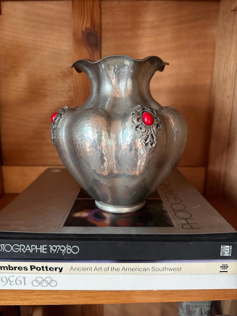 May include: A silver-toned vase with a hammered texture and a ruffled rim. The vase features ornate silver embellishments with red accents. The vase is sitting on top of a stack of books. The books include the text "PHOTOGRAPHIE 1979/80".