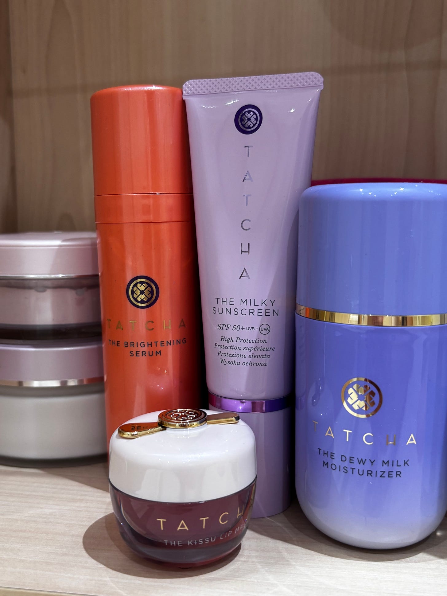 A curated lineup of Tatcha skincare on a wooden shelf for The Beauty Ed.® review. The collection highlights the "Glow-First" philosophy with the orange Tatcha Violet-C Brightening Serum, the lavender Milky Sunscreen, and the light blue Tatcha The Dewy Milk Moisturizer.