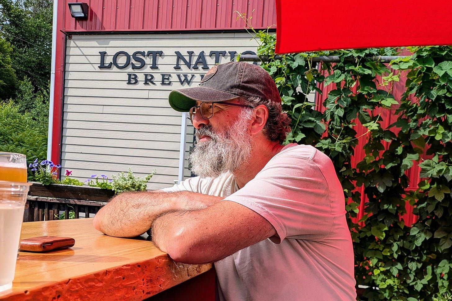 The author sits at an outdoor table at Lost Nation Brewing, arms folded, gazing into the distance in quiet contemplation. He wears a gray cap, glasses, and a pink t-shirt, his white beard catching the summer light. A craft beer and wallet rest on the wooden table beside him, with hop vines climbing a red barn wall in the background.