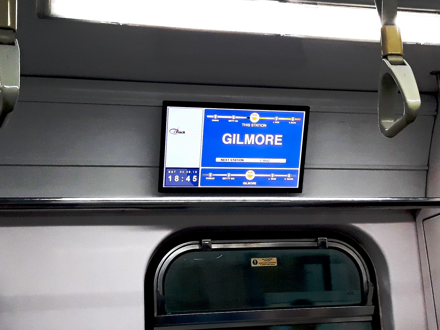 A monitor on mounted on the train's wall of LRTA Line 2. The PARDS/TUBE screen displays Gilmore, the current station, with a progress map above and below it, with the time and date shown at the bottom left