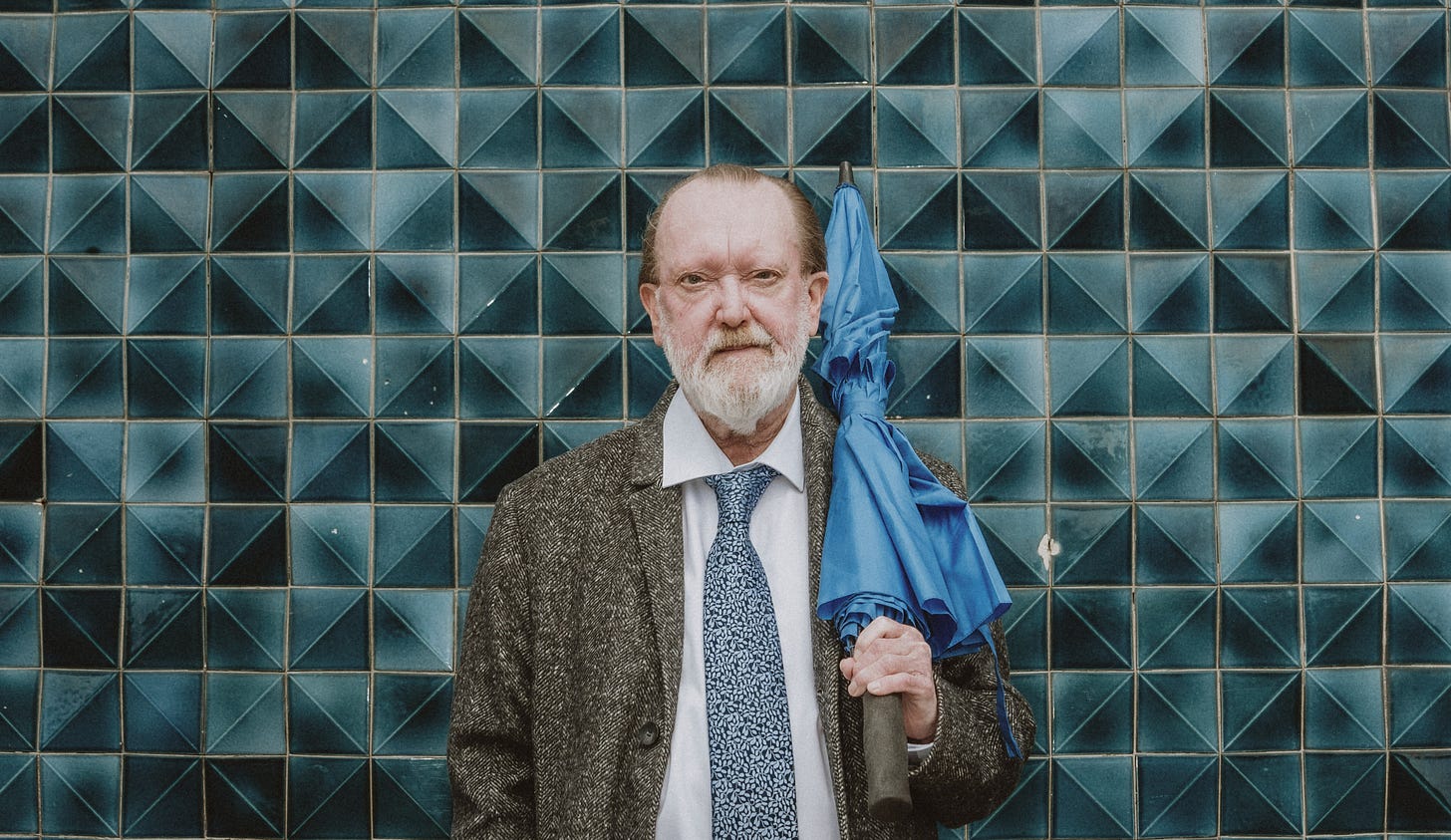 A photo of Nev Clay holding a closed umbrella, standing in front of a tiled wall.
