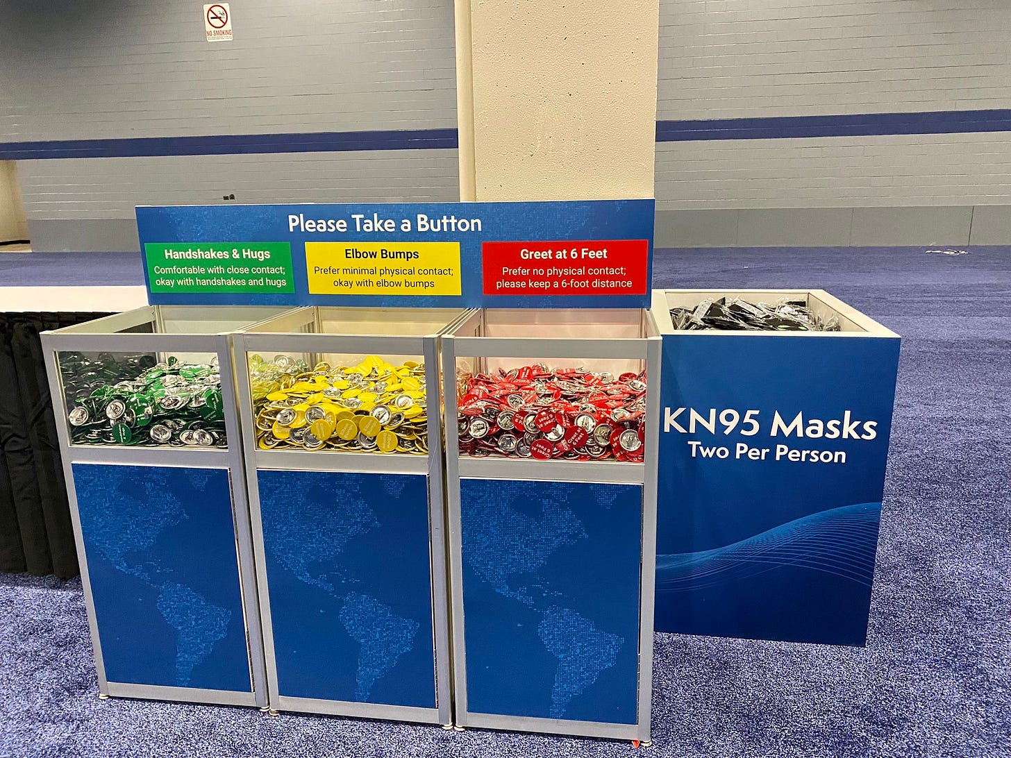 A sign says Please take a button, there are 3 bins, the first bin is labeled green and contains green buttons and the sign says Handshakes and hugs, another bin is full of yellow buttons and the yellow sign says elbow bumps, and the third bin is full of red buttons and is labeled great at 6 feet. there is a fourth bin that’s labeled KN95 masks two per person A sign says Please take a button, there are 3 bins, the first bin is labeled green and contains green buttons and the sign says Handshakes and hugs, another bin is full of yellow buttons and the yellow sign says elbow bumps, and the third bin is full of red buttons and is labeled great at 6 feet. there is a fourth bin that’s labeled KN95 masks two per person