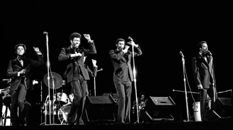 Five male musicians dressed in dark suits perform on stage in front of microphones