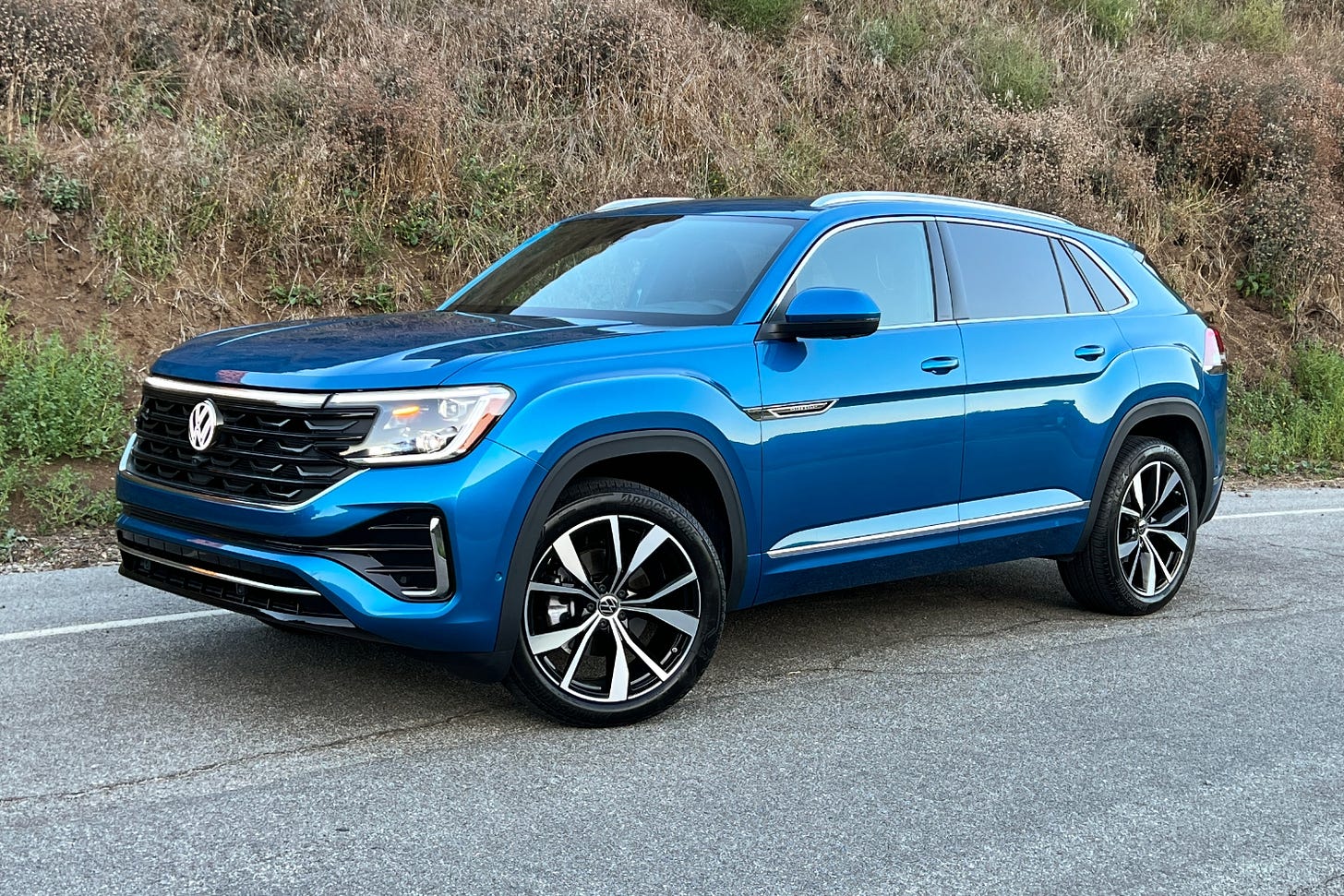 A blue 2024 Volkswagen Atlas Cross Sport parked on the side of a road with a brush-covered hillside behind it.