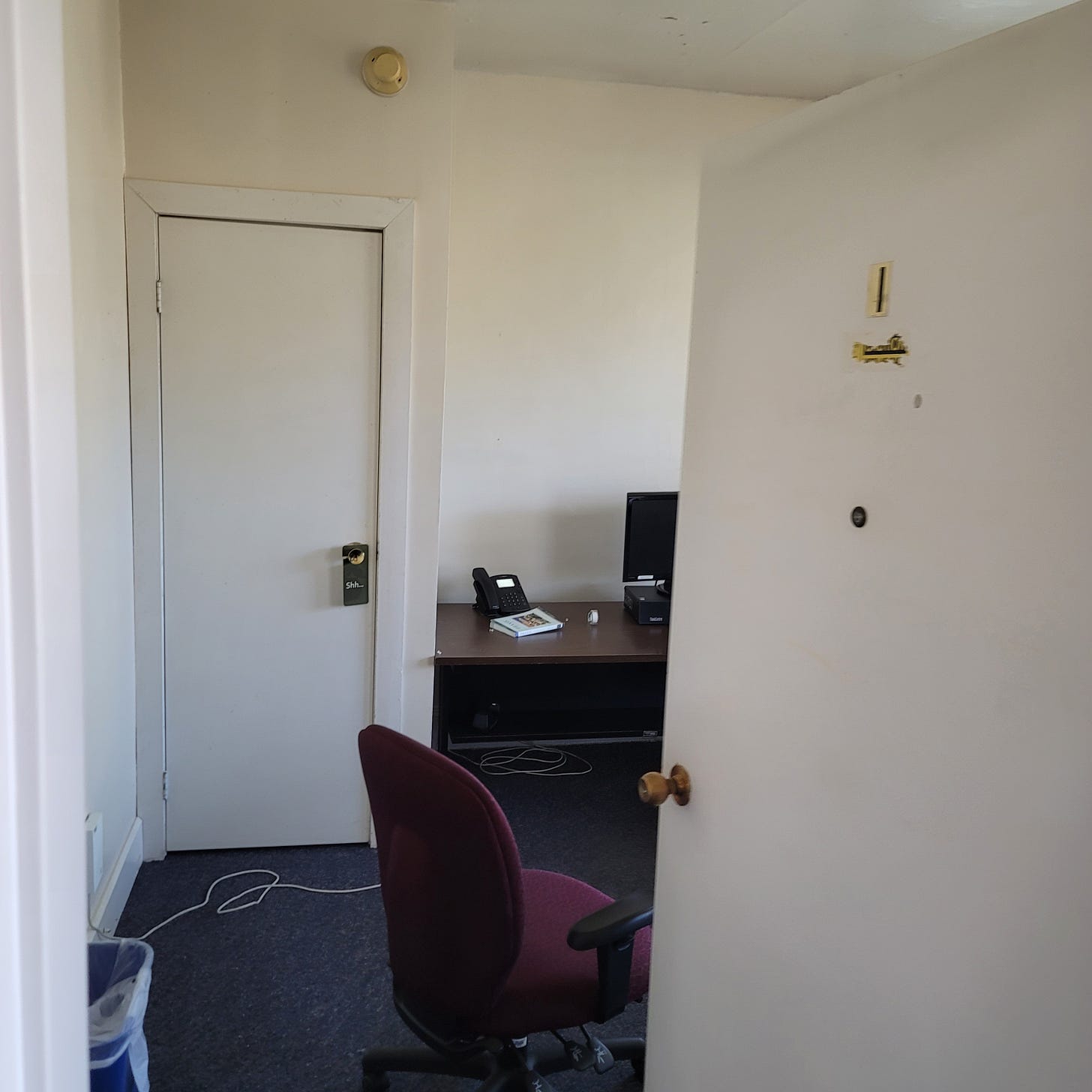 A photo of an office with the door ajar. There is an office chair and an empty room. The left side of a desk is pictured, with nothing on it except a phone, a course catalogue and an old computer.