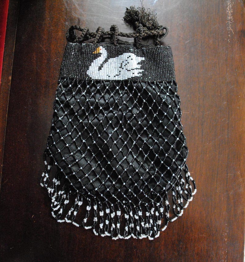 May include: Vintage black beaded evening bag with a white swan design. The bag features a mesh-like construction with black and white beads, and a fringe of black and white beads at the bottom.  The bag has a drawstring closure and a dark tassel.  The swan is embroidered in white beads on a black background.  It is a mesh style beaded purse.