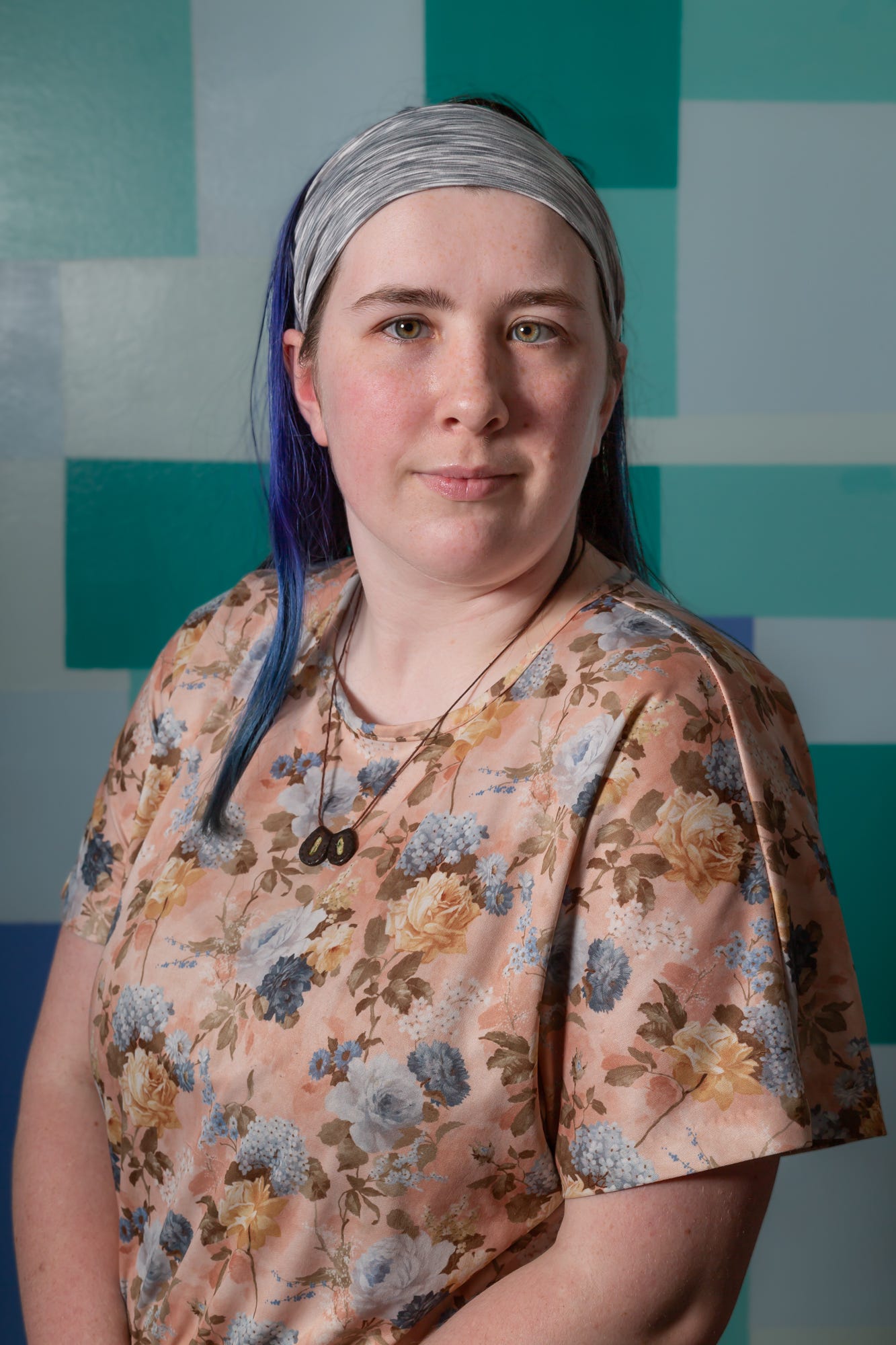 Portrait of 20 year old, white skinned woman. She is looking at the camera. She has hazel eyes and dark brown and blue hair, pulled back with a gray headband. She is wearing a pale pink floral dress.