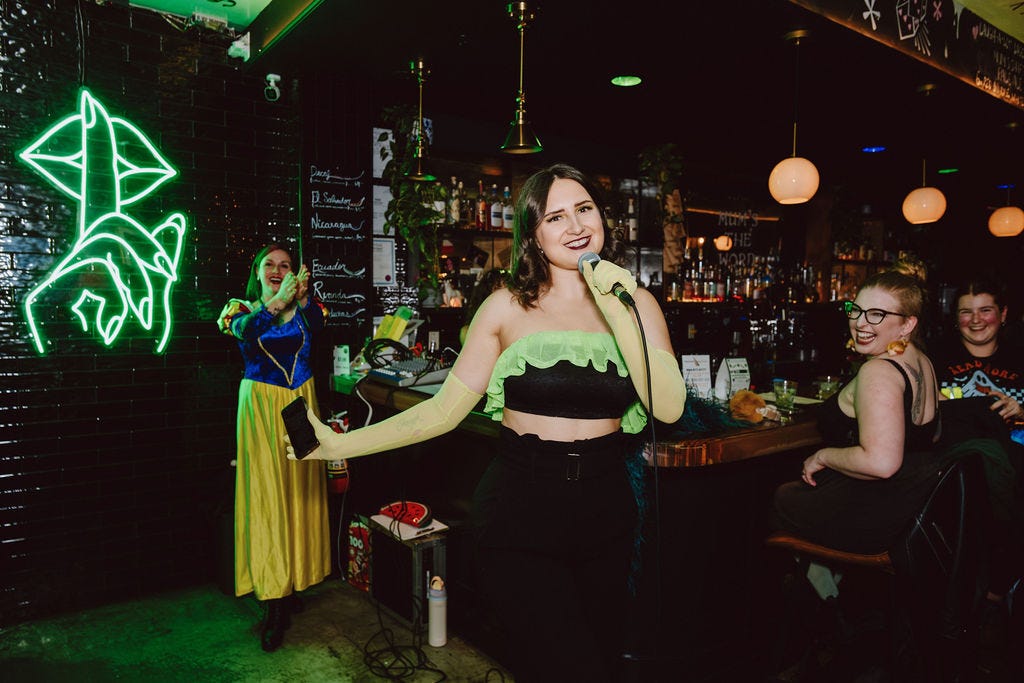 Halena Lou, a white woman wearing an apple green and black outfit, talks on a microphone smiling while people clap behind her. She is in a bar, hosting a burlesque show.