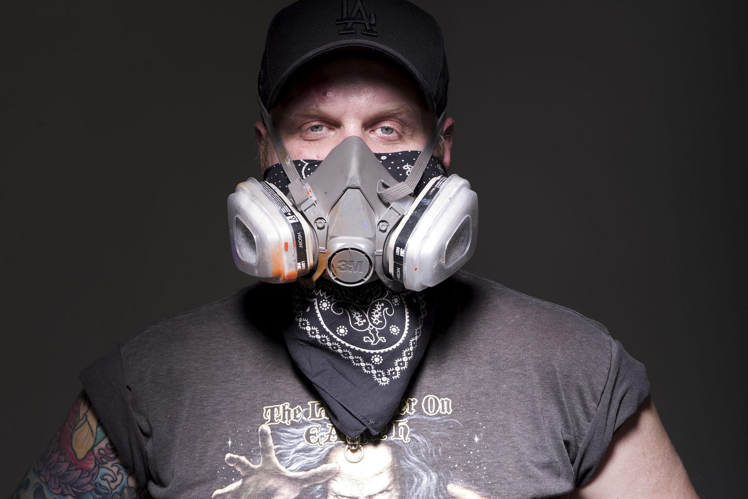 Jeremy is a white man wearing a grey respirator mask and a black bandana, wearing a black cap and a gray graphic T-shirt with rolled up sleeves with a tattooed right arm, standing against a dark background. There is orange spray paint on the front of the respirator, and his bandana is under the mask as well, covering his nose and mouth. Jeremy is staring straight into the camera. Jeremy is a white man wearing a grey respirator mask and a black bandana, wearing a black cap and a gray graphic T-shirt with rolled up sleeves with a tattooed right arm, standing against a dark background. There is orange spray paint on the front of the respirator, and his bandana is under the mask as well, covering his nose and mouth. Jeremy is staring straight into the camera.