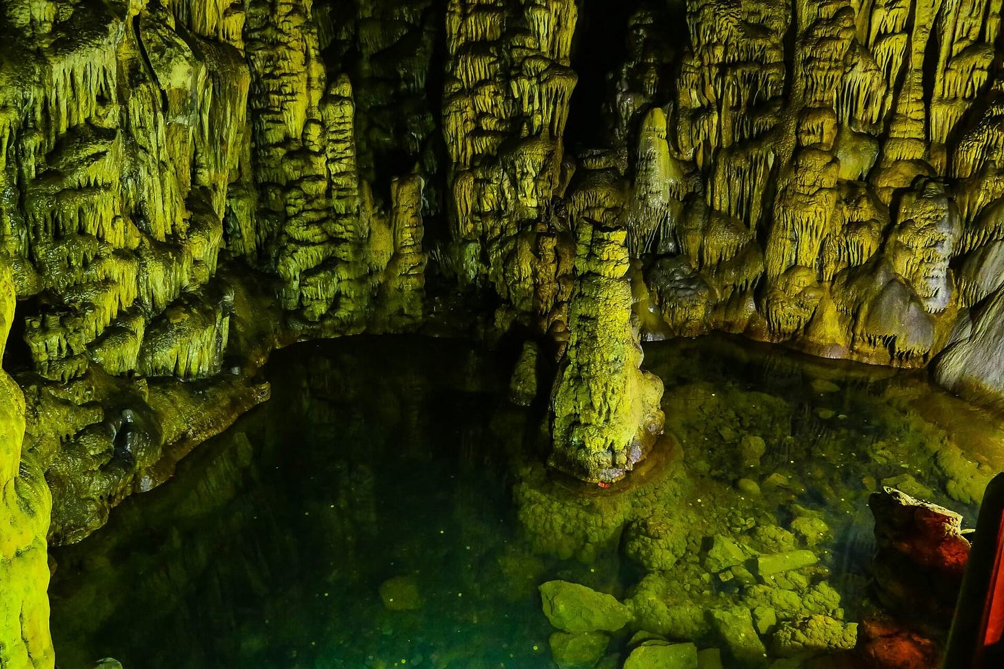 Foto do interior de uma caverna, mostrando várias estalactites e estalagmites, formações rochosas que se acumulam, como se derretessem lentamente, em tons esverdeados.