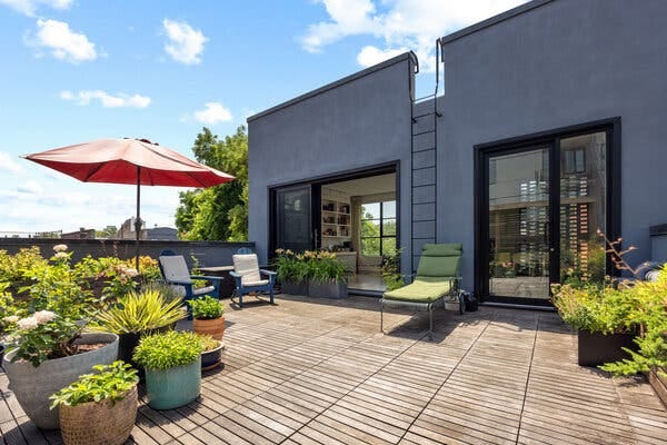 On a rooftop terrace are several potted plants and a red umbrella near a couple of chairs.