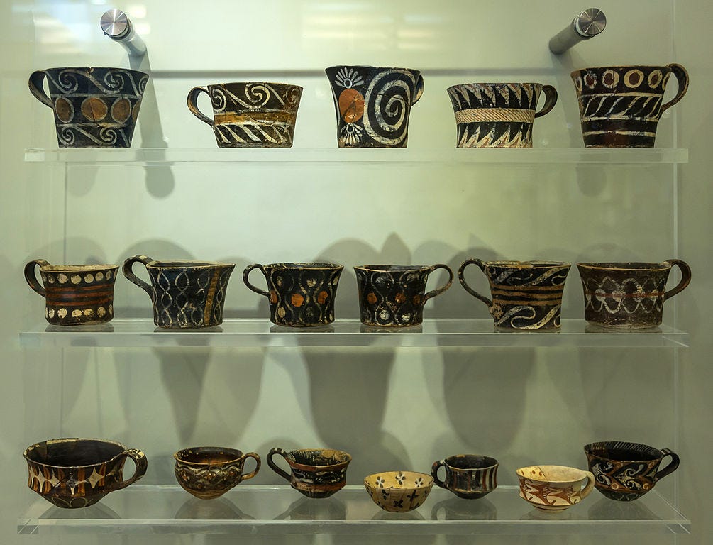 A museum display of three plexiglass shelves full of Minoan loop-handled cups, all decorated in bold designs in black, white, and red.
