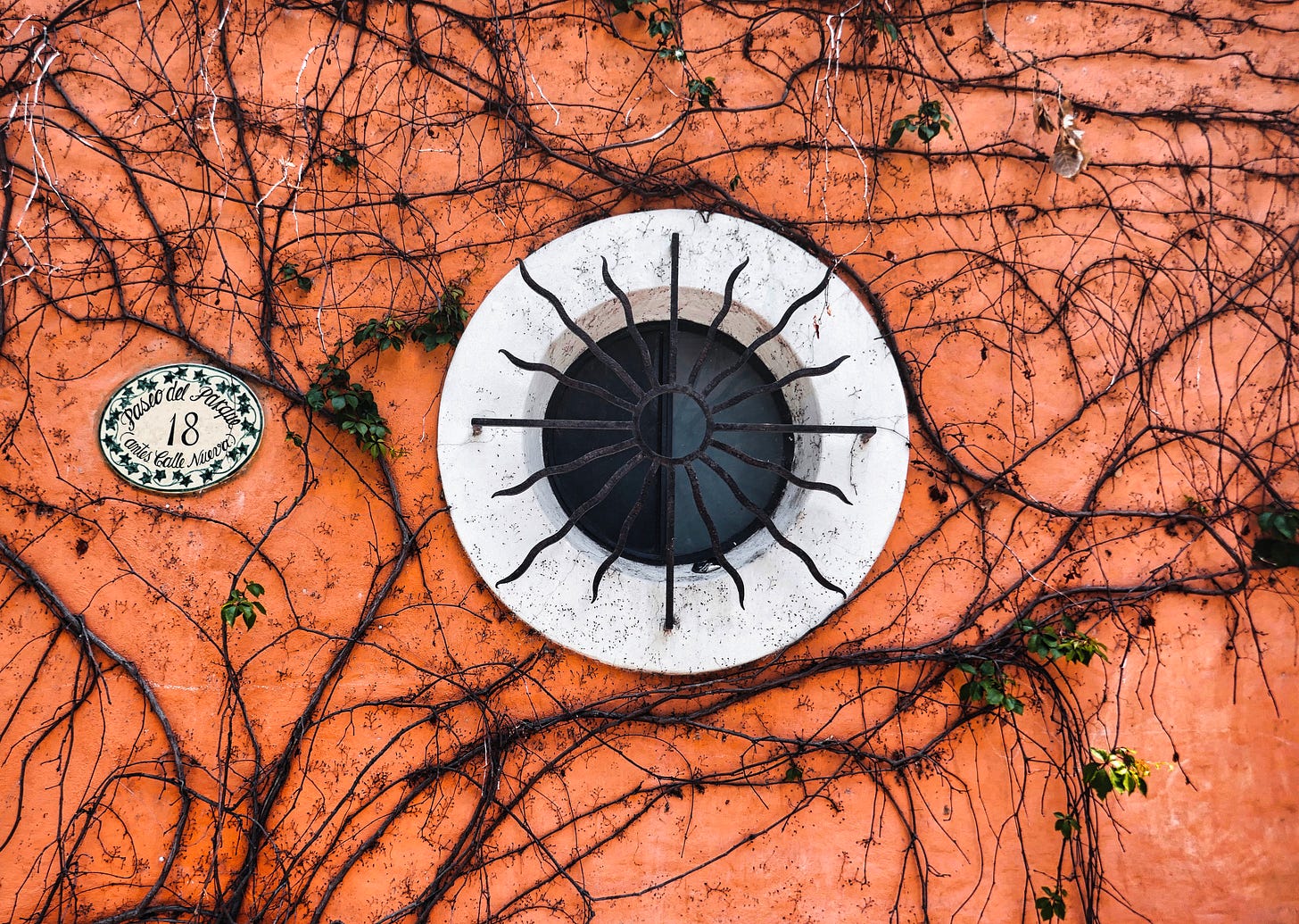 ￼a small round window with a white trim and dead vines running across the orange stucco and the street number 18 on a small plaque to the left of the window￼