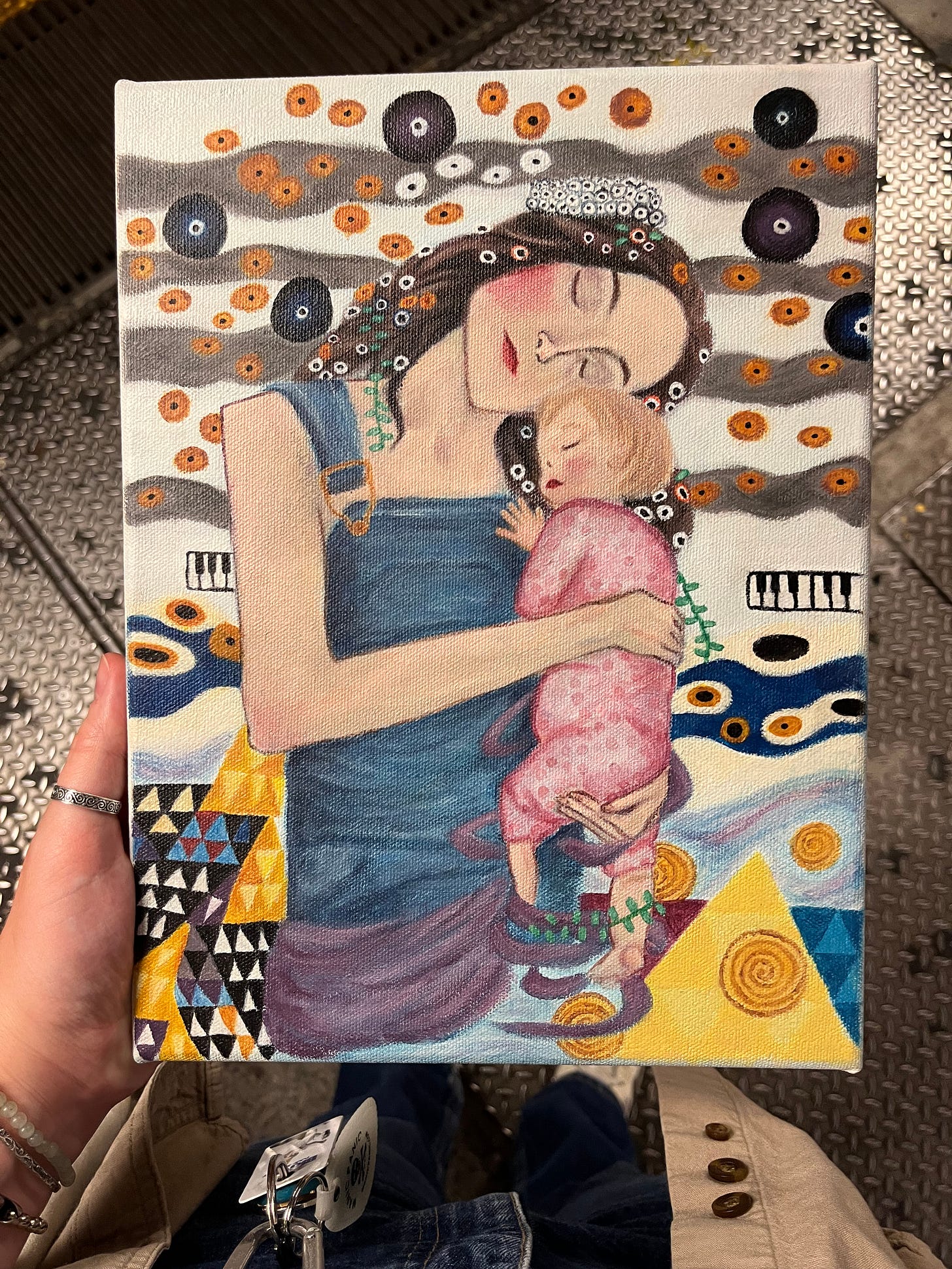 Perspective photo of someone holding up a painted canvas. The painting is a recreation of the mother and child in Klimt's The Three Ages of Woman, but the mother in this painting wears overalls and the child wears a pink onesie.