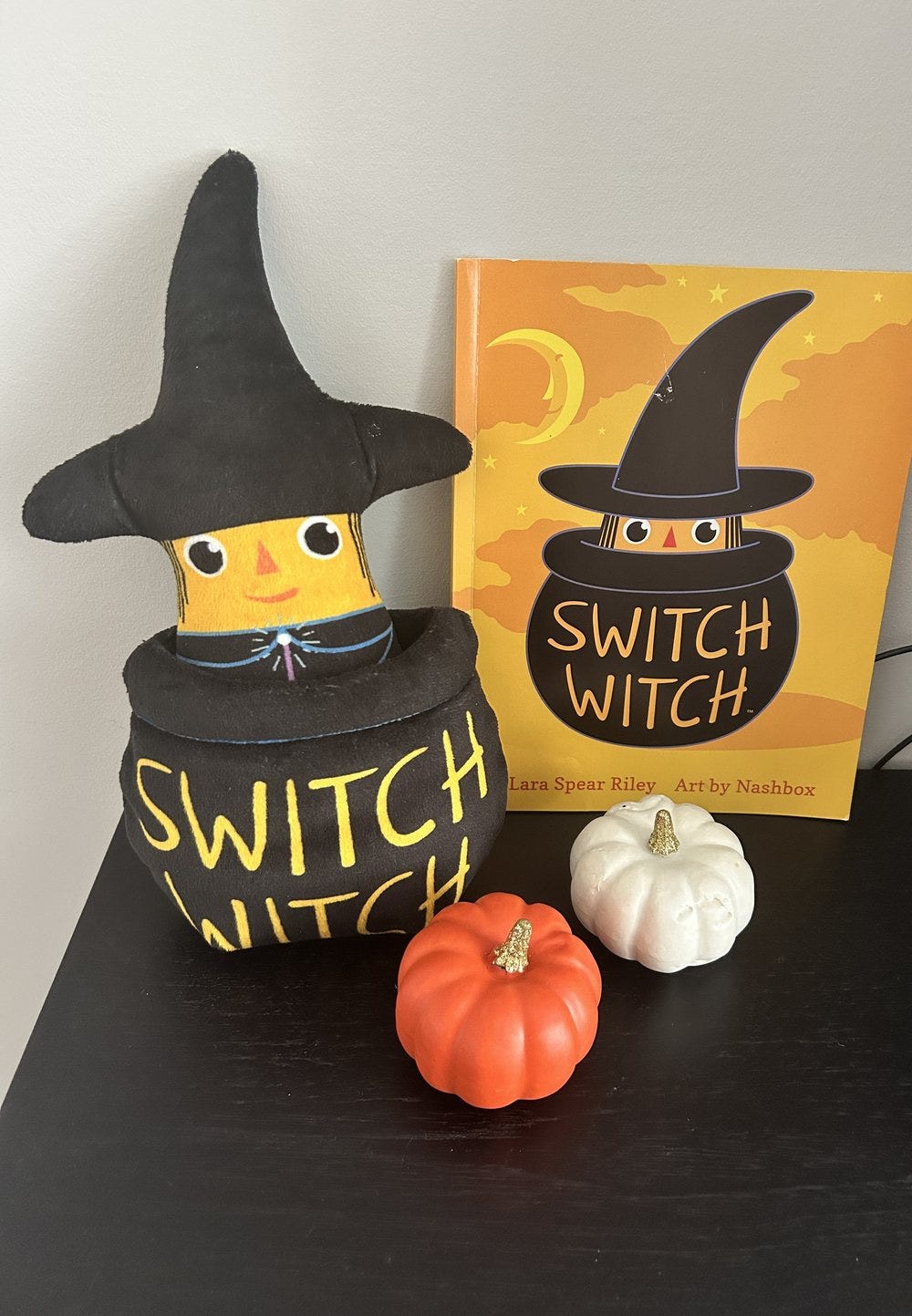 A small witch doll standing on a kitchen counter next to two small pumpkins and a storybook. A small witch doll standing on a kitchen counter next to two small pumpkins and a storybook.