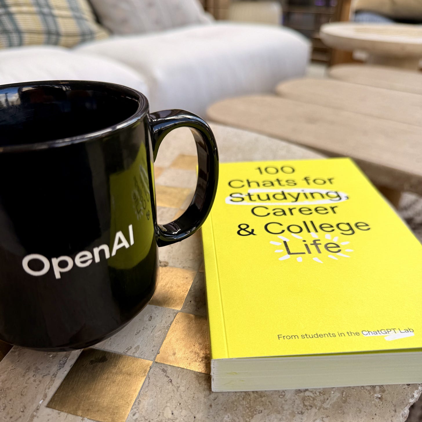 A black coffee mug with the OpenAI logo sits on a light-colored table beside a bright yellow book titled 100 Chats for Studying, Career & College Life. The setting looks casual and comfortable, with a couch and soft lighting in the background. The photo captures the personal moment that inspired reflection on how people use ChatGPT — shifting from ‘prompting’ to genuine conversation.