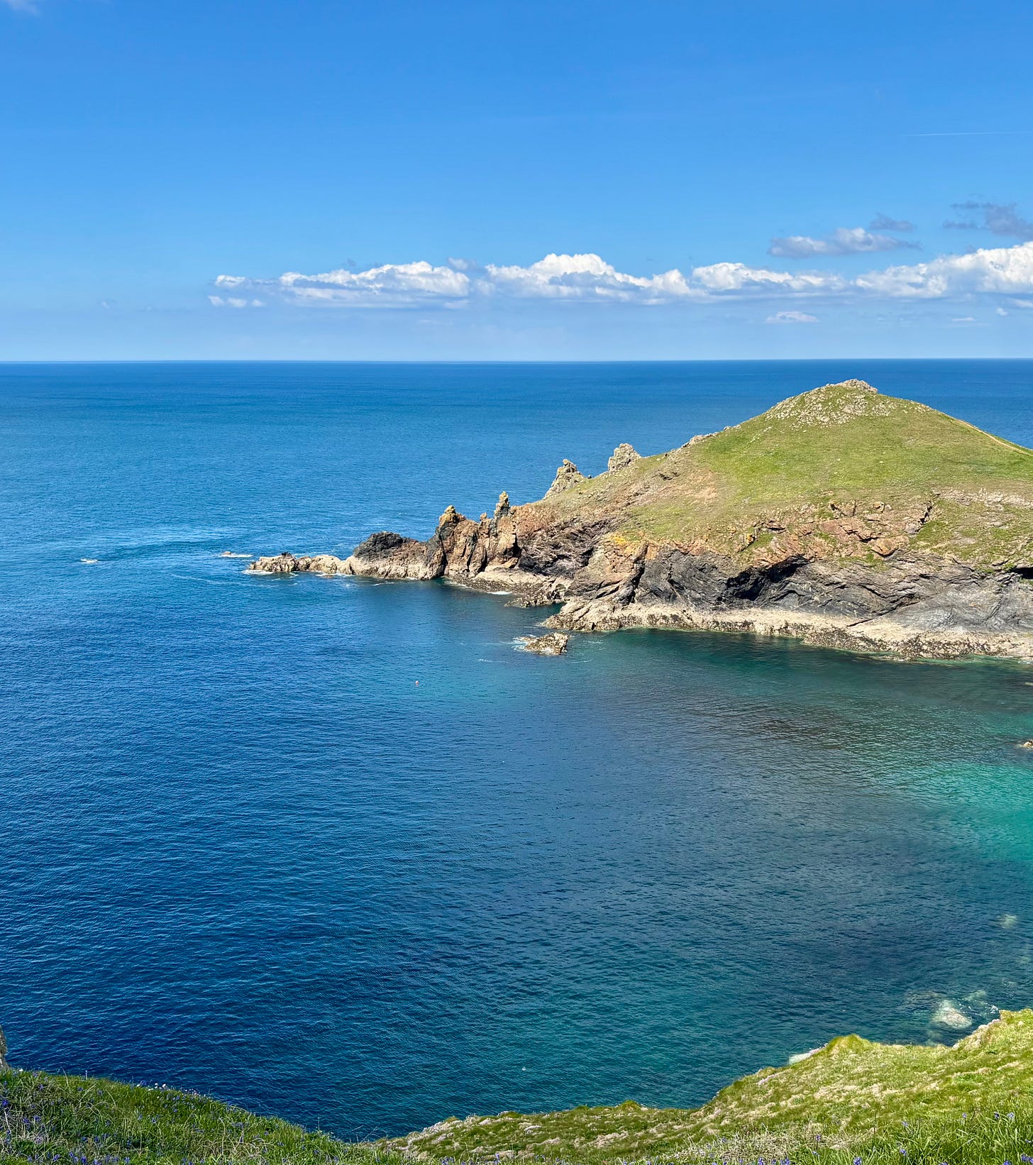 A clifftop view. The sky and sea are a vivid blue. A grassy headland with four pointy mounds lies in the middle distance. There's a tiny pale smudge in the sea at the bottom of the photo.