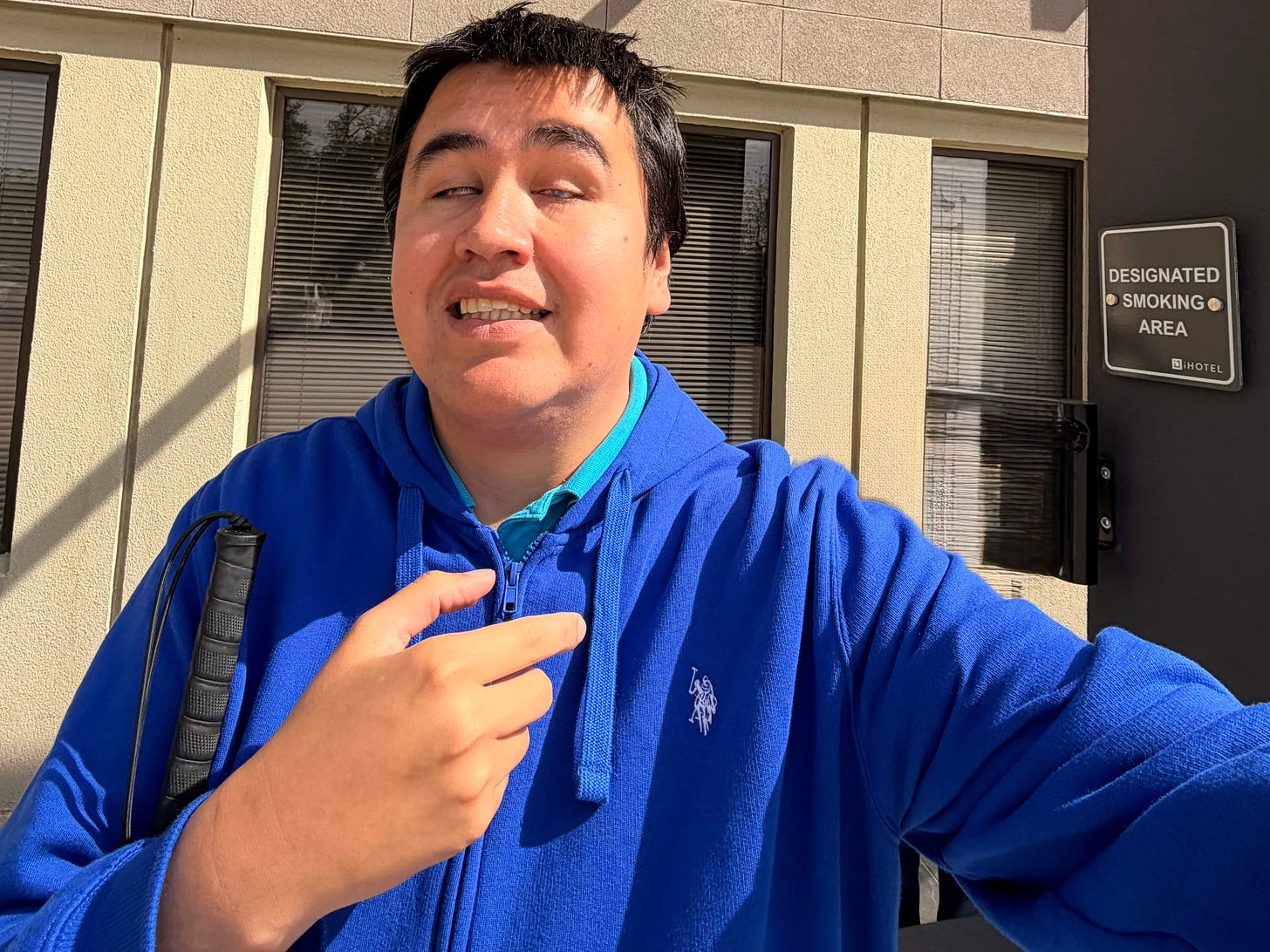 J.R. stands outside the iHOTEL in bright sunshine during a break from his one week outreach and recovery coach training program. He is wearing a bright blue hoodie, holding his white cane, and taking a selfie in front of the hotel building. A “Designated Smoking Area” sign is mounted on the wall behind him.