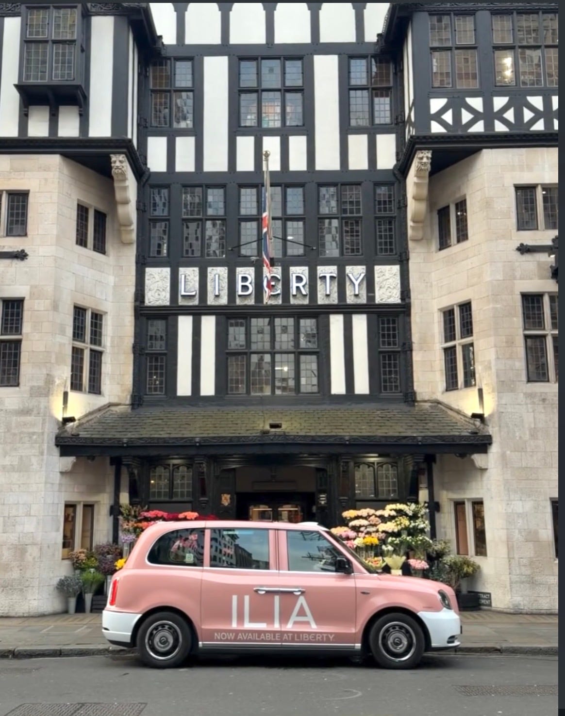 A pink ILIA Beauty branded taxi parked in front of the iconic Tudor Revival facade of Liberty London department store in the West End. A perfect blend of California beauty and British heritage.