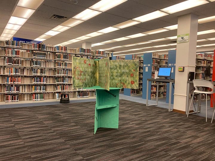 A big book with colorful pages sits on a display stand in front of a pair of windows. A painted, oversized book sits on a green display stand with library shelves visible behind it. A man stands next to an oversized book that is painted in red and green.