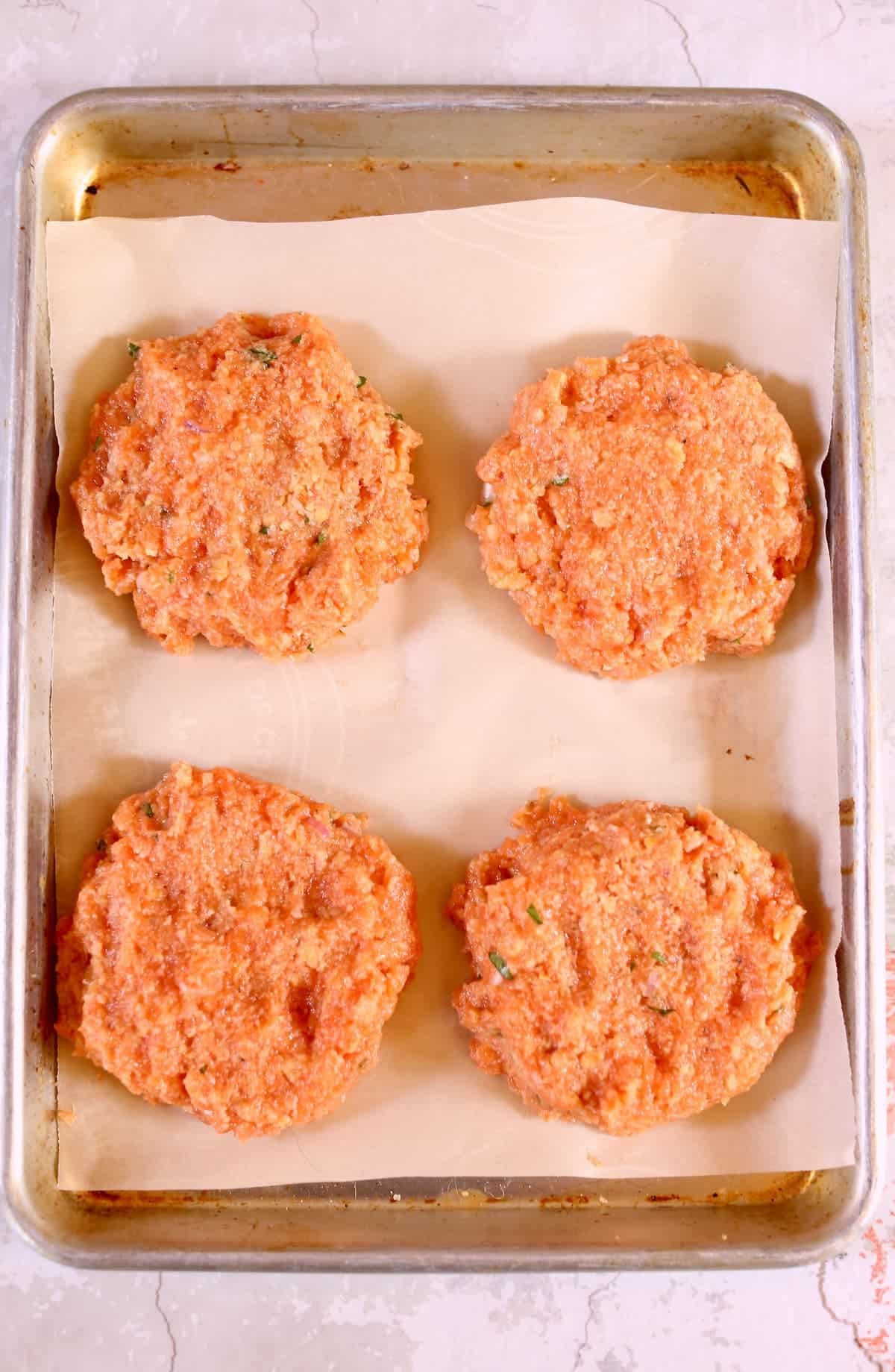 four raw salmon patties on a baking sheet.