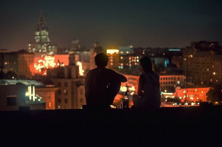 This may contain: two people sitting on top of a roof looking at the city lights in the distance