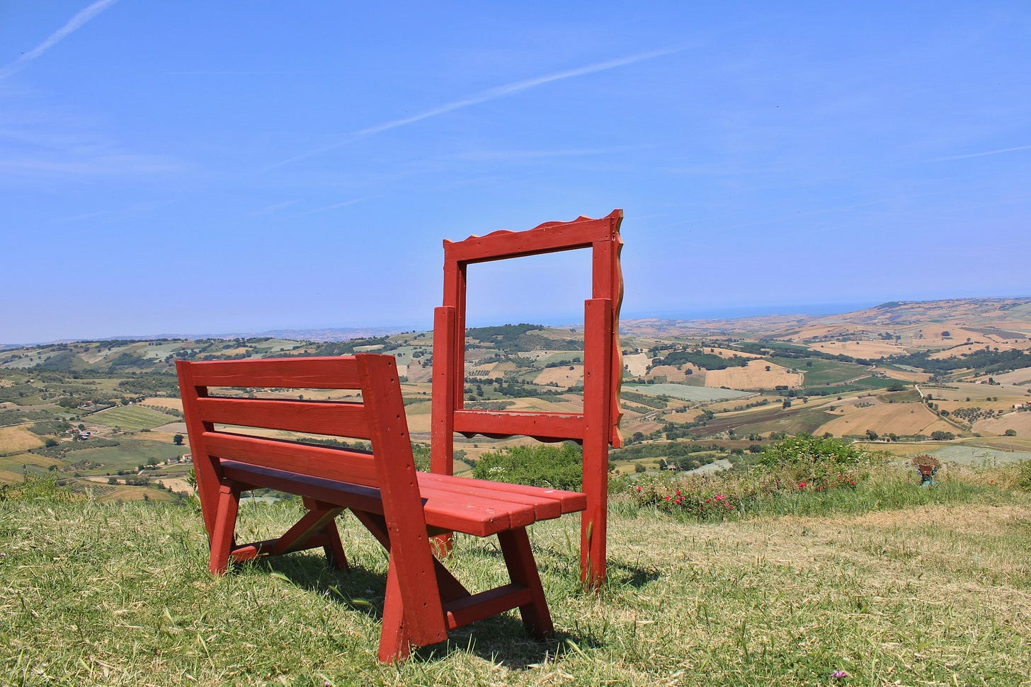 A red bench, sitting in front of a red frame, through which we can see a landscape with a patchwork of fields, houses, and farms.