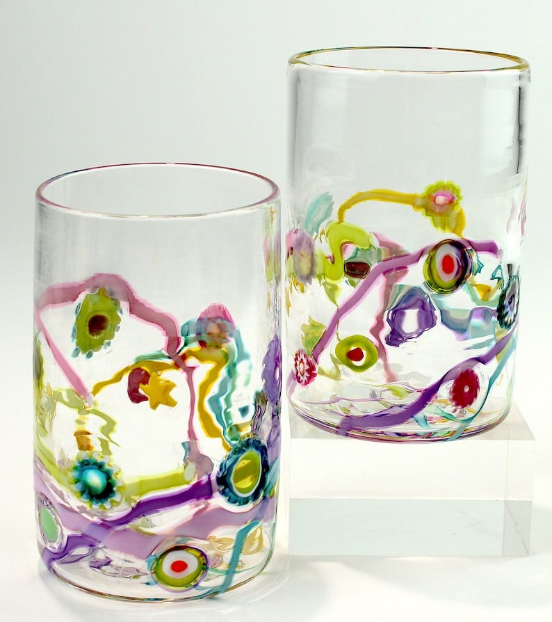 May include: Two clear glass tumblers with purple, pink, yellow, green, and blue painted designs. The tumblers are on a white surface.