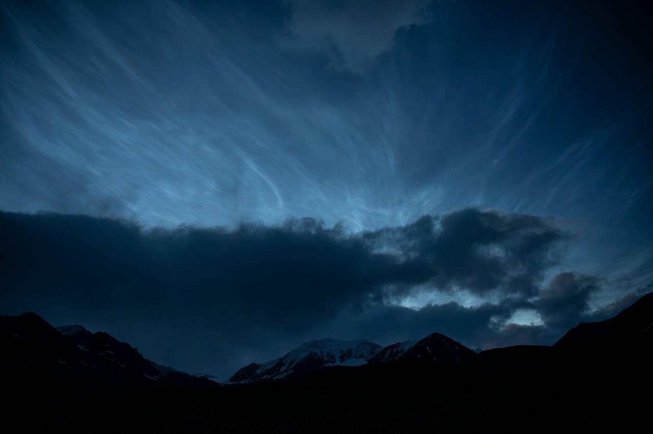 High in a dark sky, electric-blue noctilucent clouds spread in fine wavy filaments above a band of darker low cloud. Along the bottom of the frame, black mountain silhouettes show patches of snow near their summits.