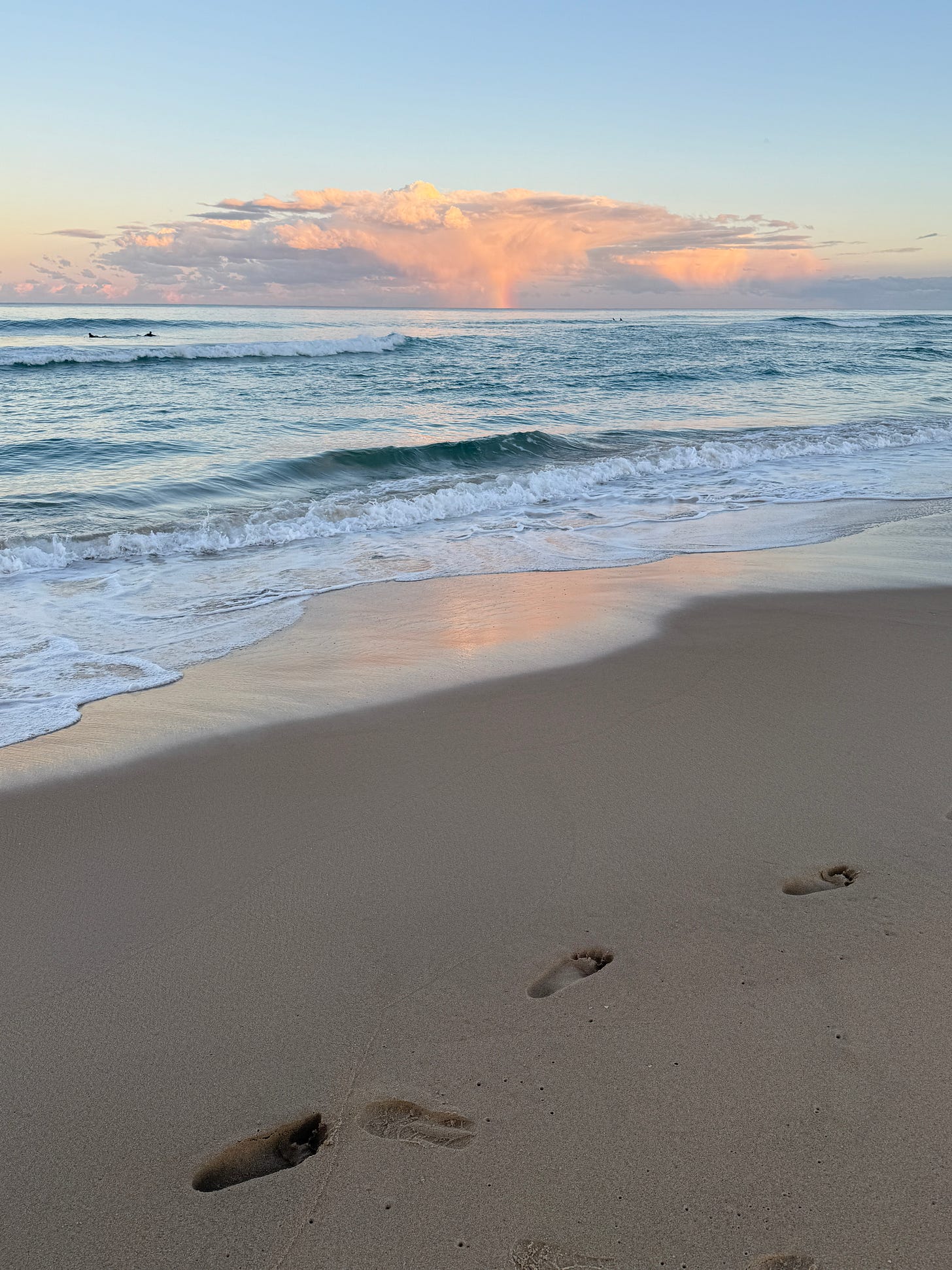 Sunshine beach, four surfers in the water, sunset, a large cloud on the horizon turning pink, a small rainbow in the middle of the cloud, footprints in the sand