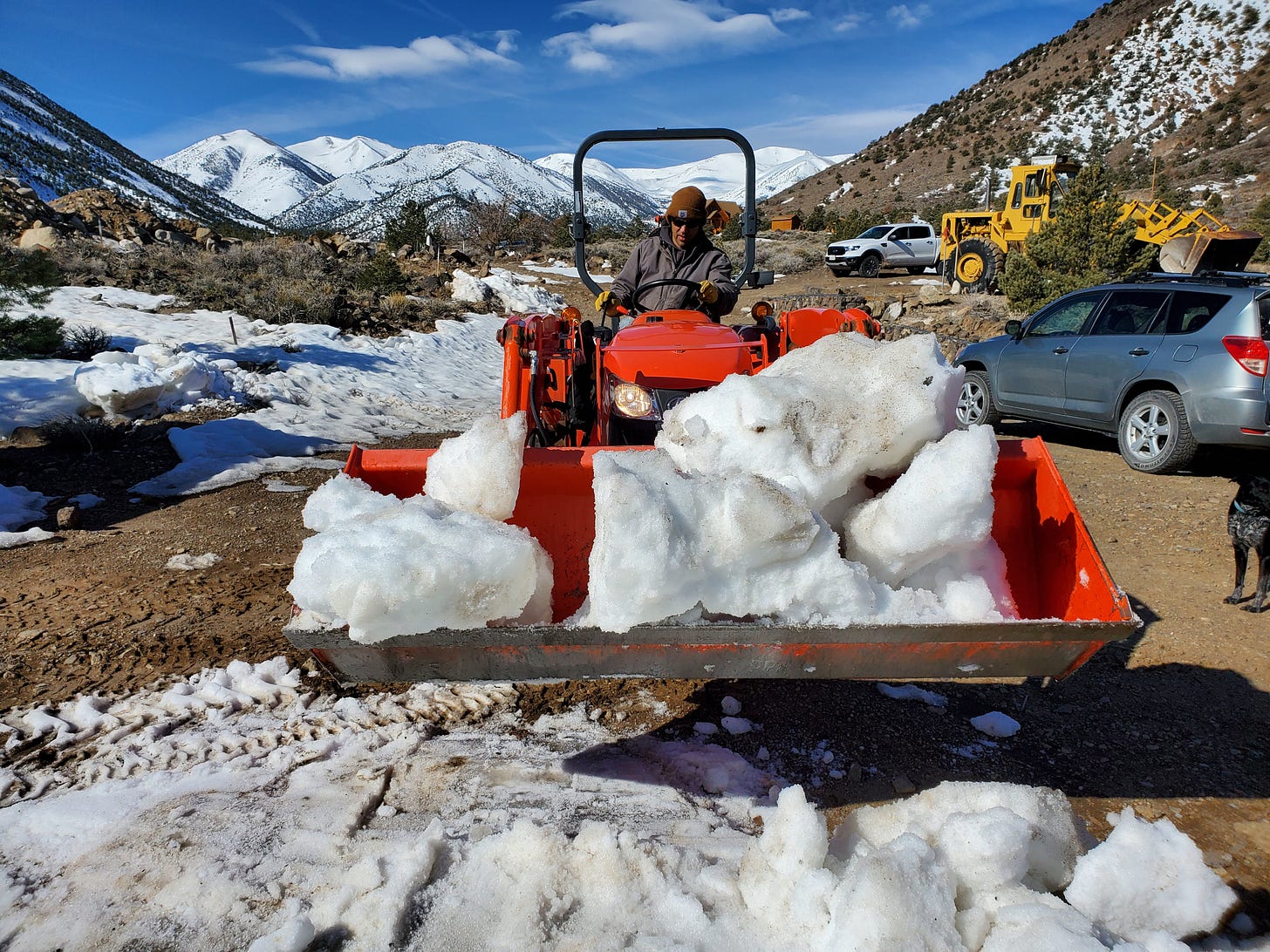 Eric driving an orange Kubota tractor. It's bucket is full of snow. White Mountains in the distance.