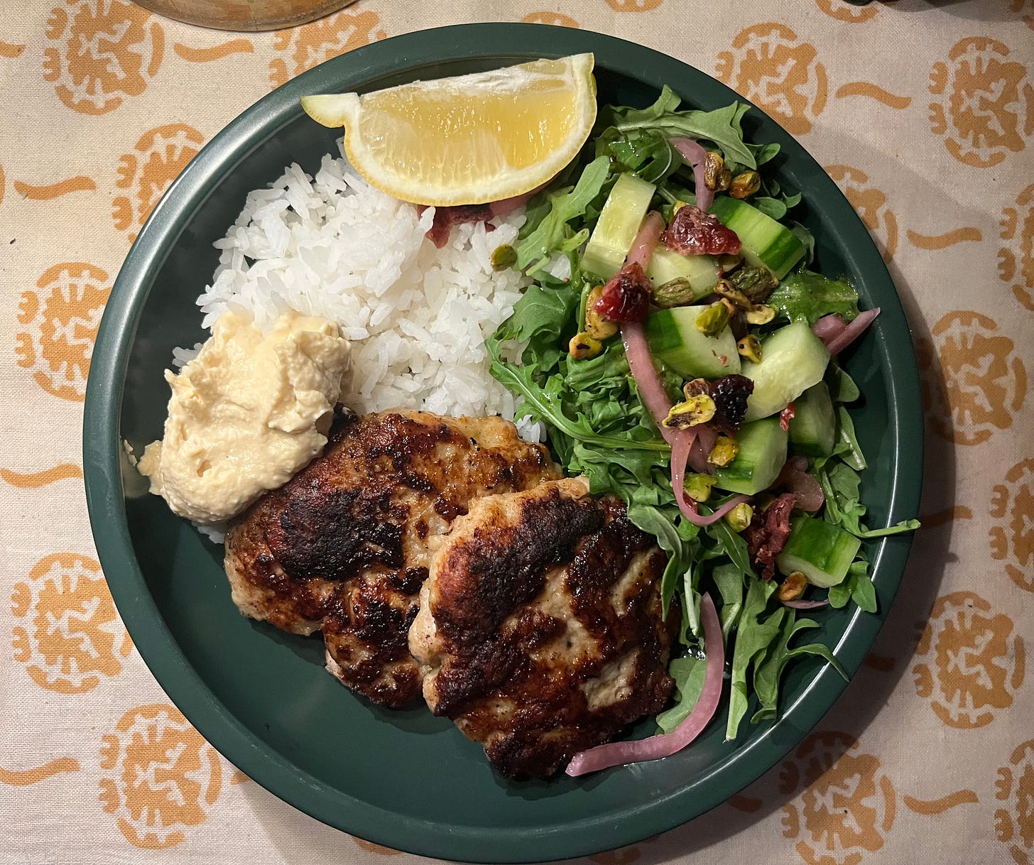 Green blat on a tan and yellow cat-printed cloth, featuring jasmine rice, hummus, lemon wedge, arugula salad with pistachios, cucumbers, and dried cranberries, pickled red onions, and two pan fried chicken cutlets. 