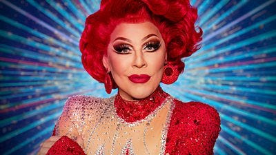 A head and shoulders shot of La Voix, a drag performer in a red wig and glittering red dress, with red earrings and  a bold statement red lip.