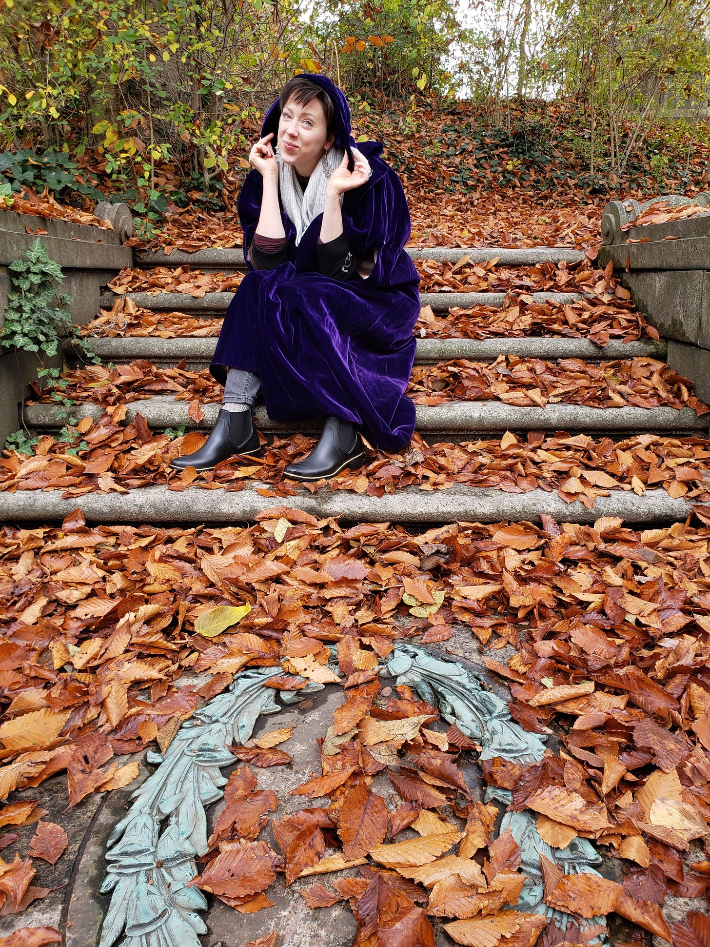 A photograph of Sam sitting on concrete steps surrounded by brown leaves and green ivy. Sam is a white woman and is wearing a violet velvet cloak and black ankle boots. There is a large, flat, pale blue ornamental wreath on the step beyond her feet.