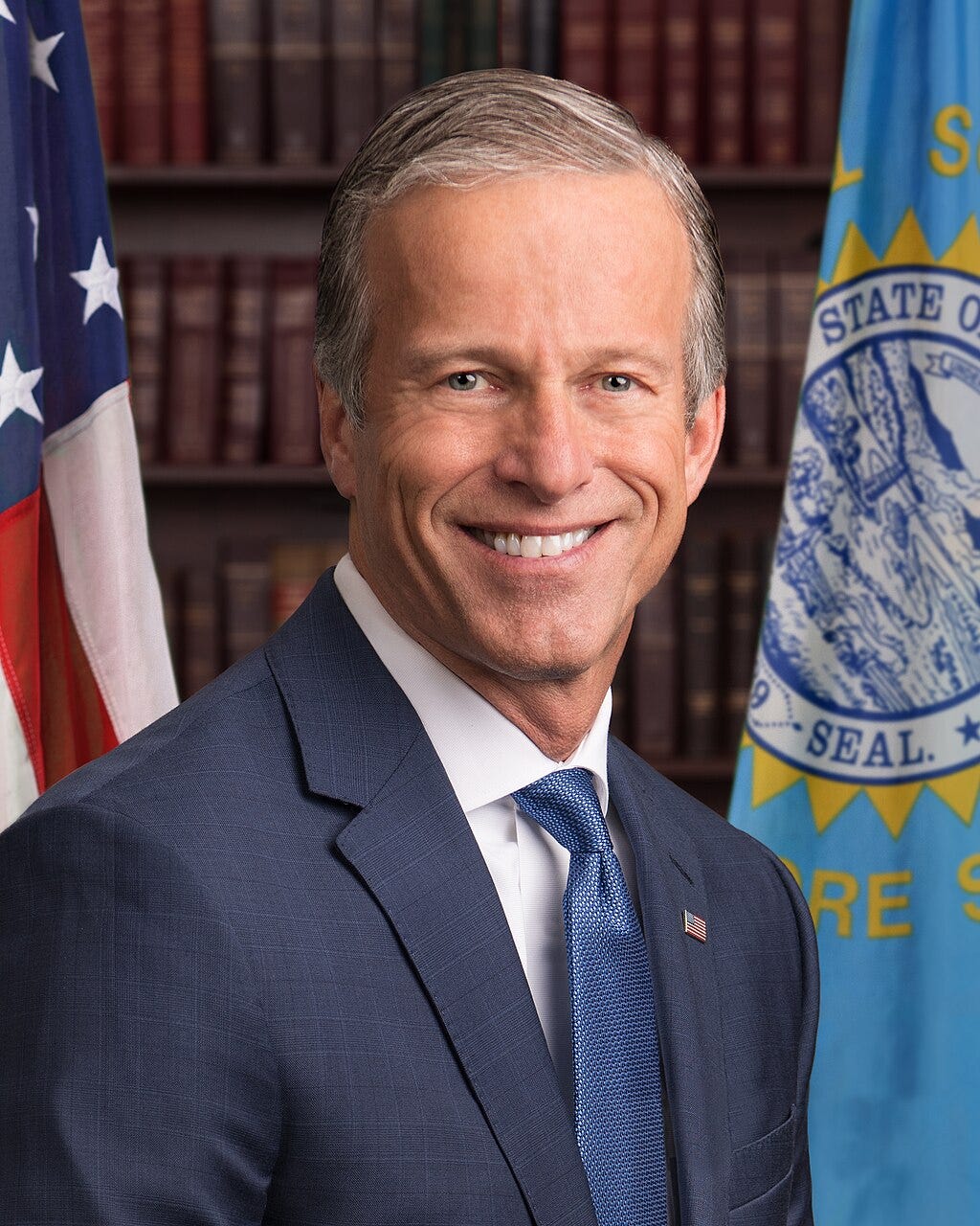 John Thune official senate portrait; turned 3/4 shoulders, face straight on smiling; suit, flag pin, blue tie, bookshelves behind him, along with American flag and state flag