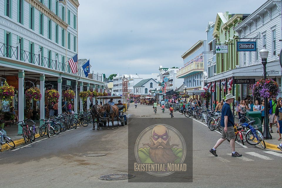  This is a wide-angle photograph of a bustling street in a town, likely on Mackinac Island, given the presence of a horse-drawn carriage and numerous bicycles. The street is unpaved and lined on both sides by multi-story buildings with covered porches and balconies. There are shops and restaurants, some with signs like "Alford's." Many people are visible on the sidewalks and in the street, either walking, riding bicycles, or riding in the carriages. Large, hanging flower baskets adorn the porches of some of the buildings. The sky is a hazy white.