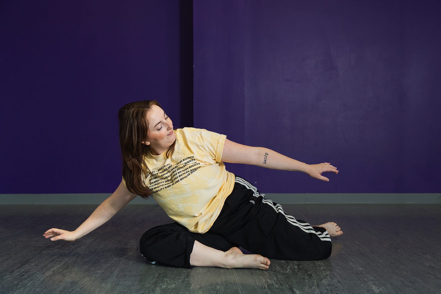 A barefoot woman in a yellow t-shirt and black triple-striped track pants sits on a grey textured floor and looks left, with both of her arms slightly raised to the side as she leans her body, against a purple corner column and wall.