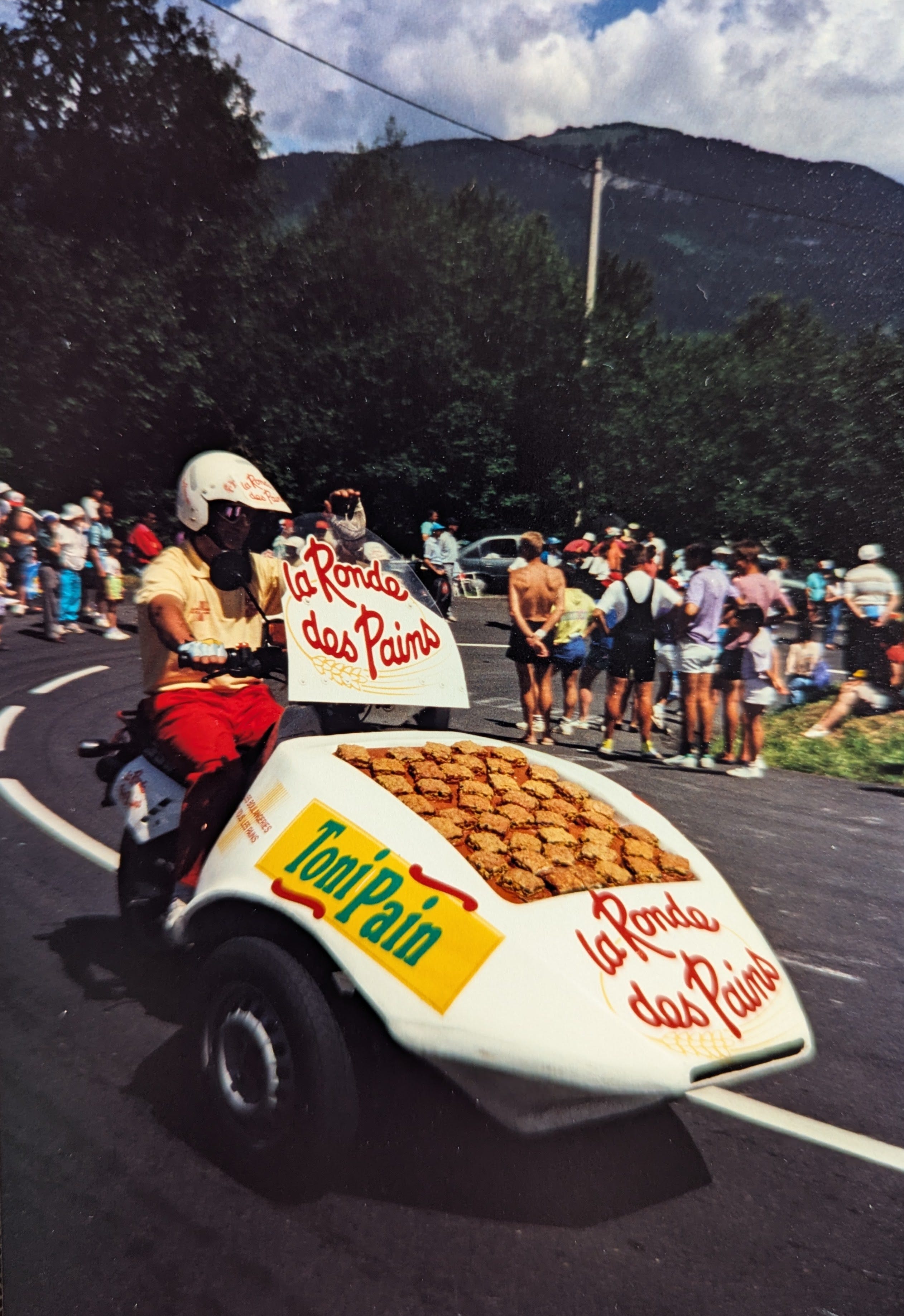 A lively outdoor scene on a mountain road in the Alps shows a person in a yellow shirt and red pants rides a tricycle - a motorcycle that has been modified with two front wheels to carry a white car-like nose. The nose is decorated with French text promoting "Campaillette" bread and "La Ronde des Pains." and on top has what appear to be biscuits or some kind of bread. Behind the motorcycle, crowds of spectators line the road. Lush green trees and dramatic mountains rise in the background under a partly cloudy sky.