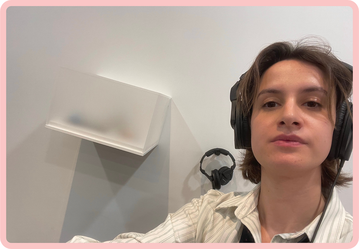 A photo of myself, Celestine, wearing a set of large black headphones, while sitting in front of an opaque glass vitrine, mounted on the wall. I am a 31 year old, white woman with dark hair in a bob, and I'm wearing a white shirt with thin green stripes. A photo of myself, Celestine, wearing a set of large black headphones, while sitting in front of an opaque glass vitrine, mounted on the wall. I am a 31 year old, white woman with dark hair in a bob, and I'm wearing a white shirt with thin green stripes.