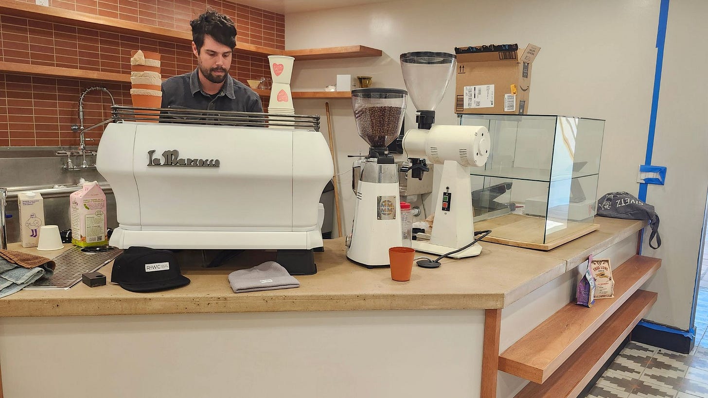 A barista works behind a white La Marzocco espresso machine against a red brick wall. The counter around him is scattered with various machine parts and coffee paraphernalia.