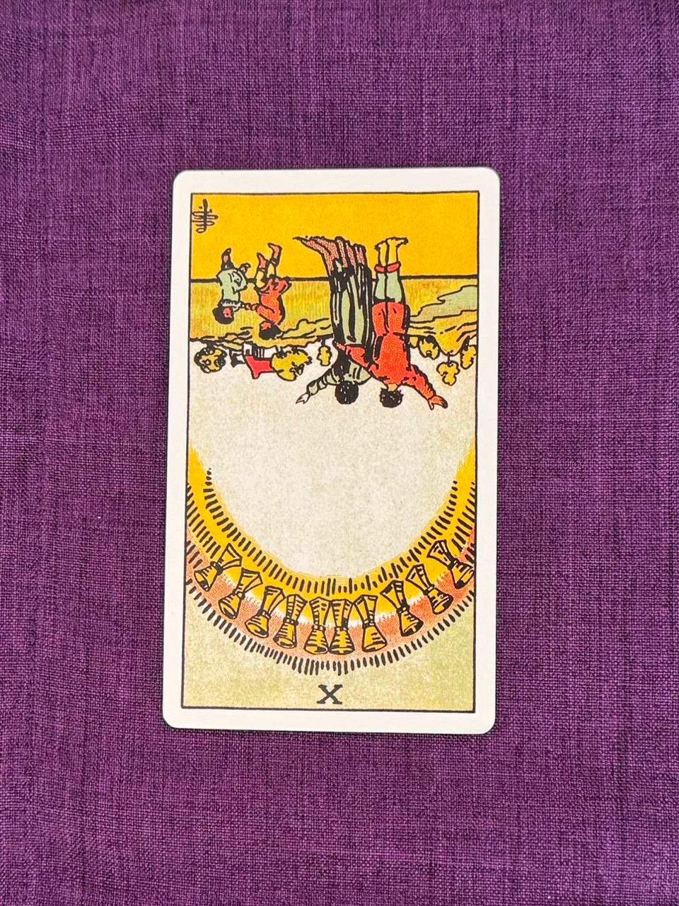 Tarot card upside down shows a family happily standing in front of the horizon. The parents face the children who are playing. There are ten cups on top, forming an arch.