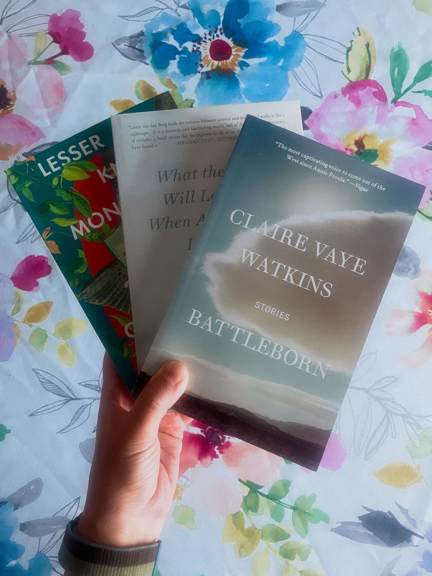 A white woman holds three paperbacks splayed like a fan. A floral tablecloth lies beneath.
