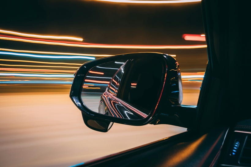 A side-view mirror of a car is in sharp focus in the center of the image, with a little of the window frame and dashboard visible below. The car window and door are reflected in the mirror, but nothing inside the car is visible. The rest of the frame shows long horizontal lines of light in gold, red, and blue, as if the picture was taken with a long-exposure lens while the car was moving past lights at night. The feeling of the photo is futuristic in a retro sort of way, alien and without any sign of humans. A side-view mirror of a car is in sharp focus in the center of the image, with a little of the window frame and dashboard visible below. The car window and door are reflected in the mirror, but nothing inside the car is visible. The rest of the frame shows long horizontal lines of light in gold, red, and blue, as if the picture was taken with a long-exposure lens while the car was moving past lights at night. The feeling of the photo is futuristic in a retro sort of way, alien and without any sign of humans.
