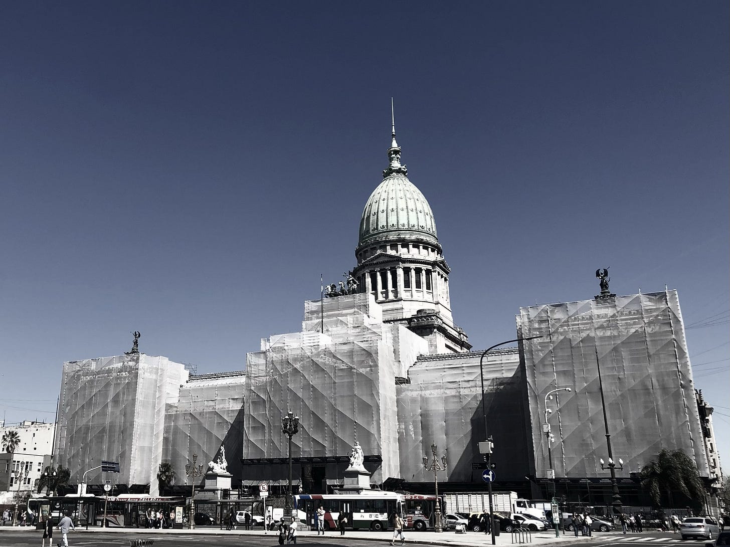 El Palacio del Congreso de la Nación, con su cúpula verde, cubierto de andamios y telas de obra blancas bajo un cielo azul, con colectivos y peatones en la avenida. El Palacio del Congreso de la Nación, con su cúpula verde, cubierto de andamios y telas de obra blancas bajo un cielo azul, con colectivos y peatones en la avenida.