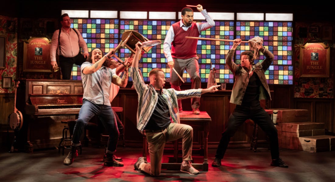 A group of five men on stage holding chairs and instruments, with a multi-coloured glass window behind it