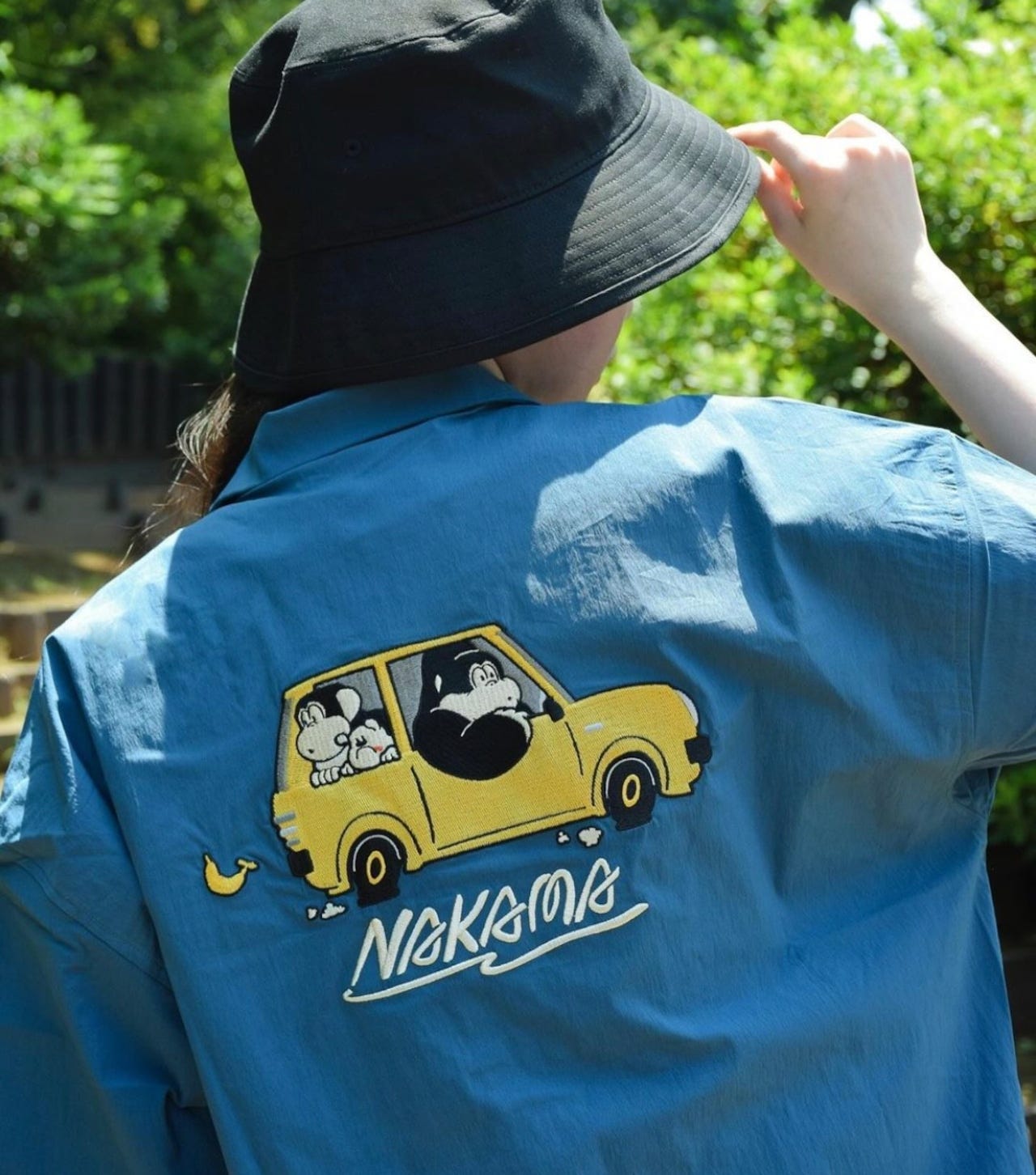 Back view of a person in a bucket hat wearing a teal-blue work shirt. The rear panel is embroidered with Joe Mountain driving a yellow hatchback while two smaller gorilla pals ride in back. Back view of a person in a bucket hat wearing a teal-blue work shirt. The rear panel is embroidered with Joe Mountain driving a yellow hatchback while two smaller gorilla pals ride in back.