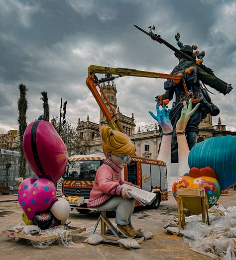 A gallery of Fallas monuments in Valencia, Spain — elaborately sculpted figures including a Charlie Chaplin figure holding butterflies, fantasy warriors, and colorful street sculptures, photographed against dramatic cloudy and blue skies in March 2026.