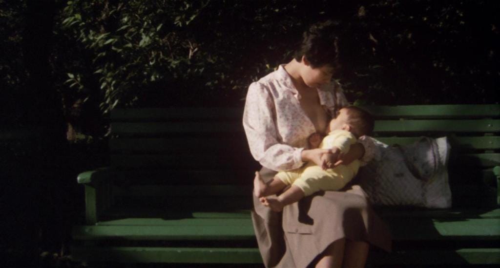 A woman sits on a green park bench, breastfeeding her baby. She wears a patterned blouse, looking down gently. Natural light and foliage surround them, evoking tranquillity.